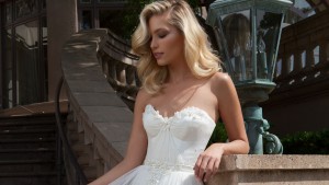Blonde woman leaning against wall by stairs. Right hand is holding the top layer of this strapless a-line bridal gown adorned with 3d detailing.