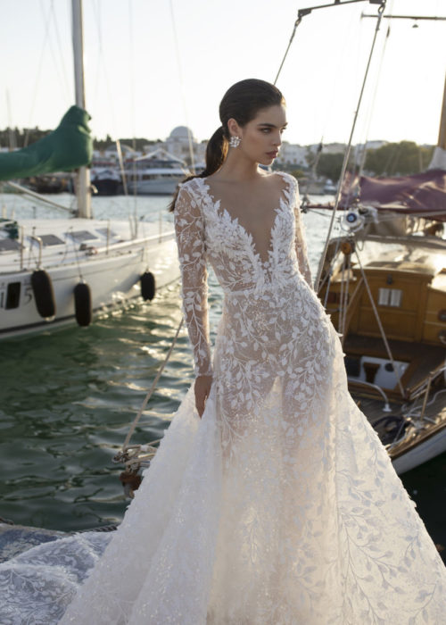 Woman standing by the waters. A-line bridal gown with an adorned plunging neckline and fitted long sleeves. This highly detailed gown showcases the body through thin lining to give a touch of sexiness to the gown.