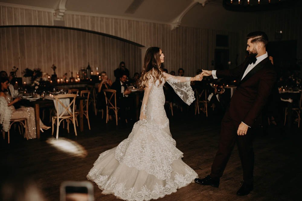 Bride to be on the dance floor with her partner. Long sleeve bell shape sleeve. Bride smiles at her partner and holds his left hand.