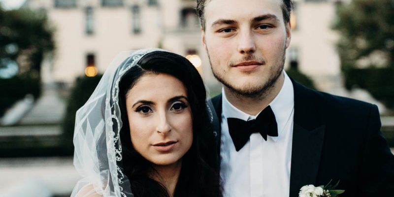 Nava adorned with is sheer veil as she stands alongside her husband Blake on their wedding day. Behind Blake is a blurred background.