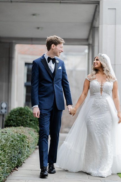 Deveen and her husband holding hands as they walk side by side. Husband is wearing a classic blue suit. Deveen is wearing the Josephine gown with an overskirt